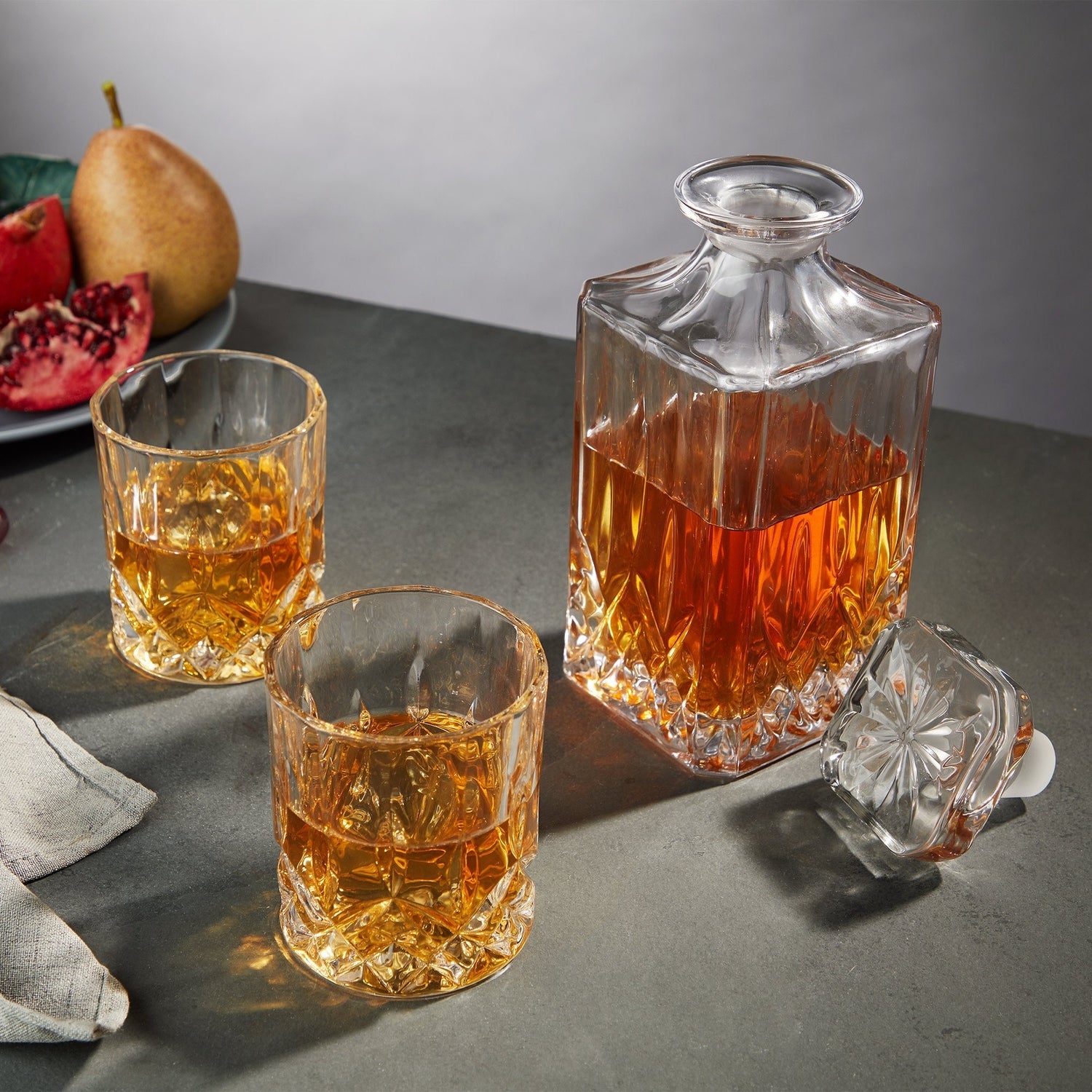 A still life photograph showcasing a square crystal decanter filled with amber liquid alongside two matching cut-glass tumblers on a dark grey tabletop, with a light brown pear and a vibrant red sliced pomegranate in the background