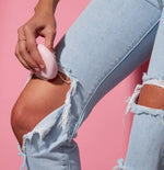 Hand holding a small pink hair removal device against a knee in ripped blue jeans