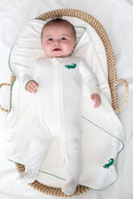 Baby lying in a woven basket wearing a white footed sleeper with green crocodile embroidery