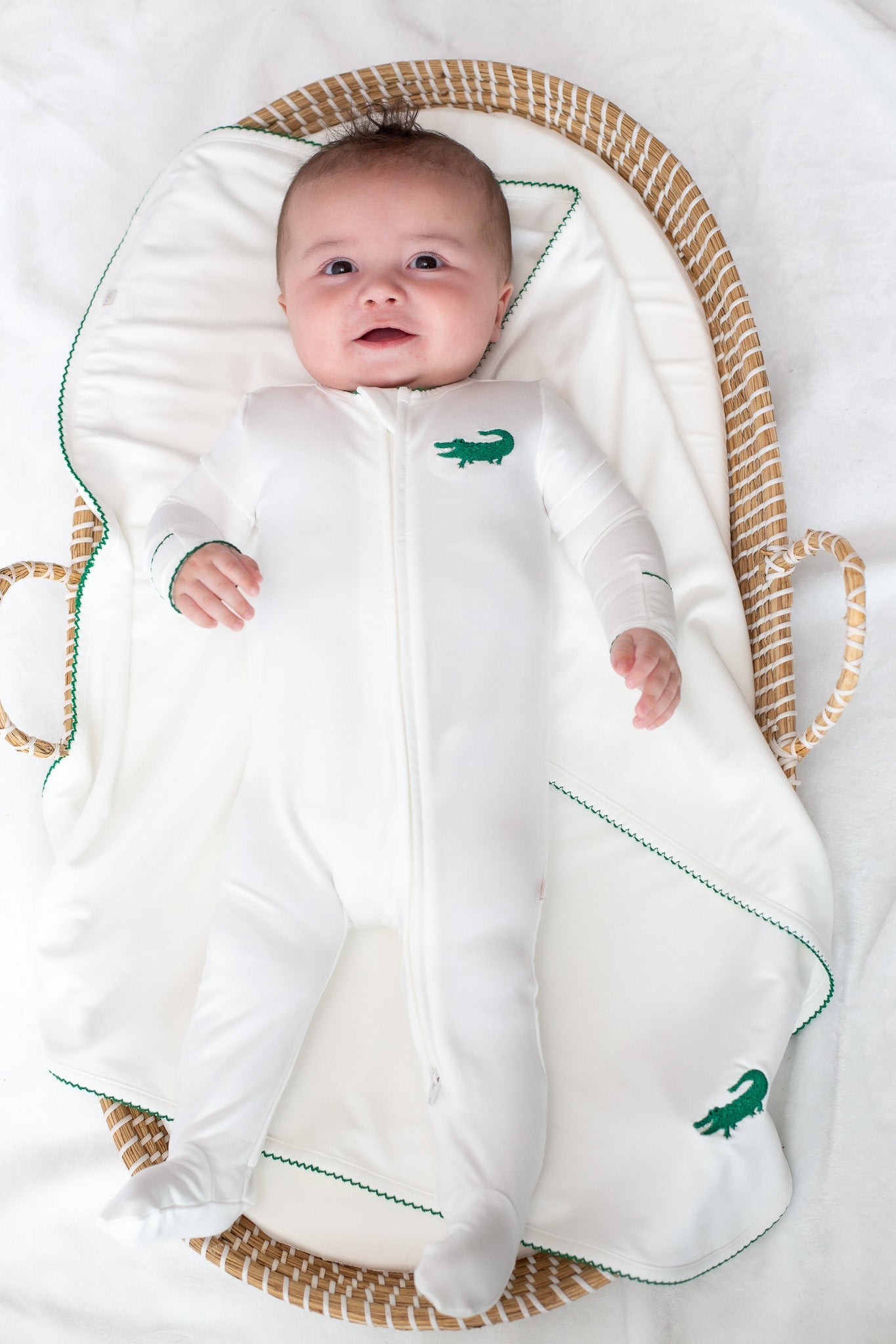 Baby lying in a woven basket wearing a white footed sleeper with green crocodile embroidery