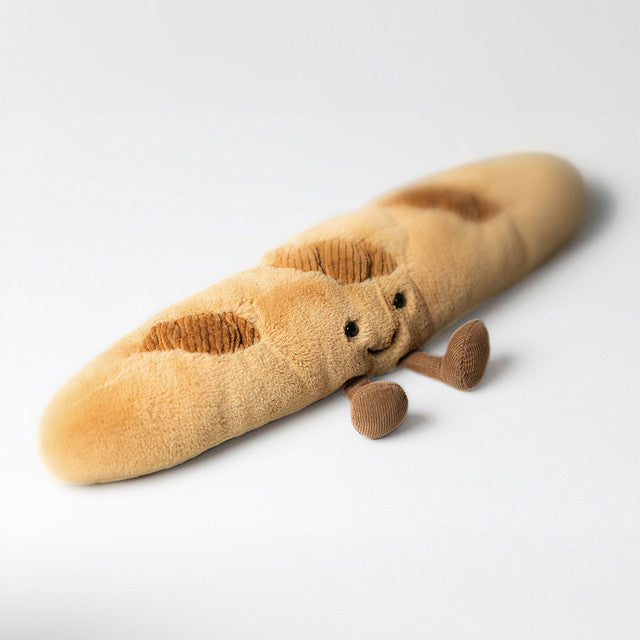 A golden brown Jellycat Amuseable Baguette plush toy with dark brown scorings, an embroidered smiling face, and small corduroy arms and legs is lying on a plain white background