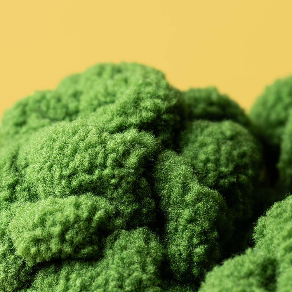 A close-up photograph featuring vibrant green fluffy textured material with soft, irregular, rounded forms suggestive of moss or miniature landscapes set against a bright yellow backdrop