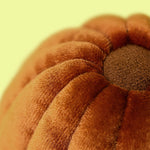Close-up photograph of a plush orange-brown velvet pumpkin pillow featuring ribbed segments and a darker brown dimpled center against a soft yellow blurred background