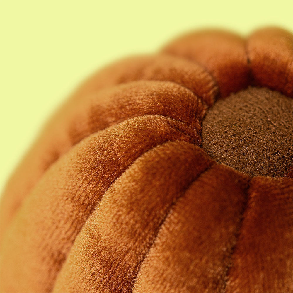 Close-up photograph of a plush orange-brown velvet pumpkin pillow featuring ribbed segments and a darker brown dimpled center against a soft yellow blurred background