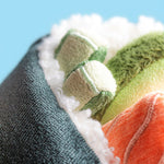 Plush toy by Jellycat featuring a macro view of a maki sushi roll with fuzzy white sherpa rice, a bright orange salmon insert, and two green cucumber slices against a solid light blue background