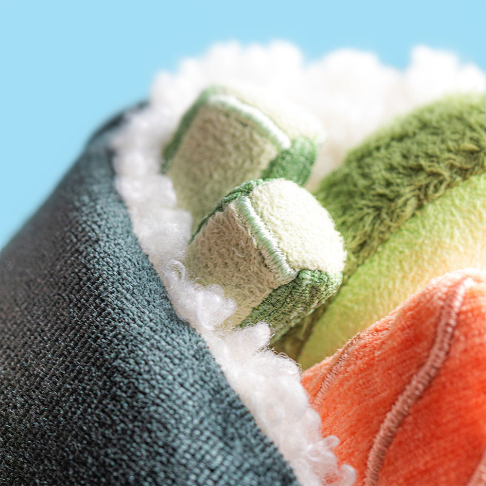 Plush toy by Jellycat featuring a macro view of a maki sushi roll with fuzzy white sherpa rice, a bright orange salmon insert, and two green cucumber slices against a solid light blue background