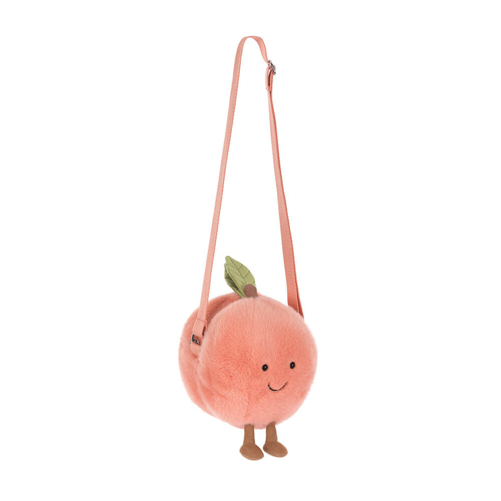 Jellycat plush crossbody bag shaped like a fuzzy light pink peach with a green leaf, cute embroidered smiling face, small brown feet, and an adjustable pink strap
