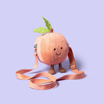 Photograph by Jellycat featuring a fuzzy peach-shaped plush bag with an embroidered smiling face, green felt leaves, and a pinkish-orange adjustable strap, on a solid lavender background