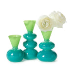 Blown glass a trio of modern decorative vases in glossy teal and lime green with stacked globular bases and funnel necks one holding two delicate white ranunculus flowers against a stark white background