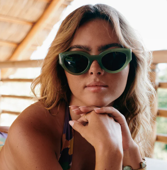 Close-up portrait of a young woman with blonde wavy hair wearing olive green cat-eye sunglasses and a vibrant patterned bikini top, her hands clasped, against a rustic straw-paneled ceiling
