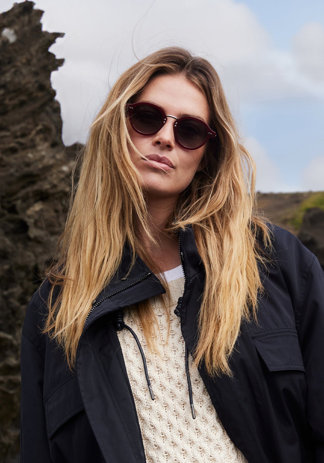 Digital photography of a woman with long blonde hair and maroon round sunglasses wearing a navy utility jacket over a cream cable knit sweater against a backdrop of dark jagged rocks