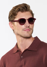 Close-up studio portrait of a man with wavy brown hair and light stubble wearing stylish maroon round sunglasses with silver rivets and a textured dark red polo shirt against a crisp white background
