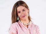 Photograph of a smiling young woman against a white studio background wearing a soft pink cardigan, a chunky white beaded link necklace, and unique triangular beaded earrings with red floral patterns