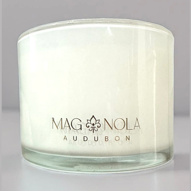 Product photography of a Magnolia Audubon white candle in a clear glass jar, featuring elegant gold branding with a prominent fleur-de-lis, referencing New Orleans