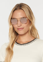 A smiling blonde woman wearing hexagonal tortoise-shell sunglasses with pink lenses and a gold double bridge, dressed in an off-white ribbed shirt with black decorative stitching around the collar
