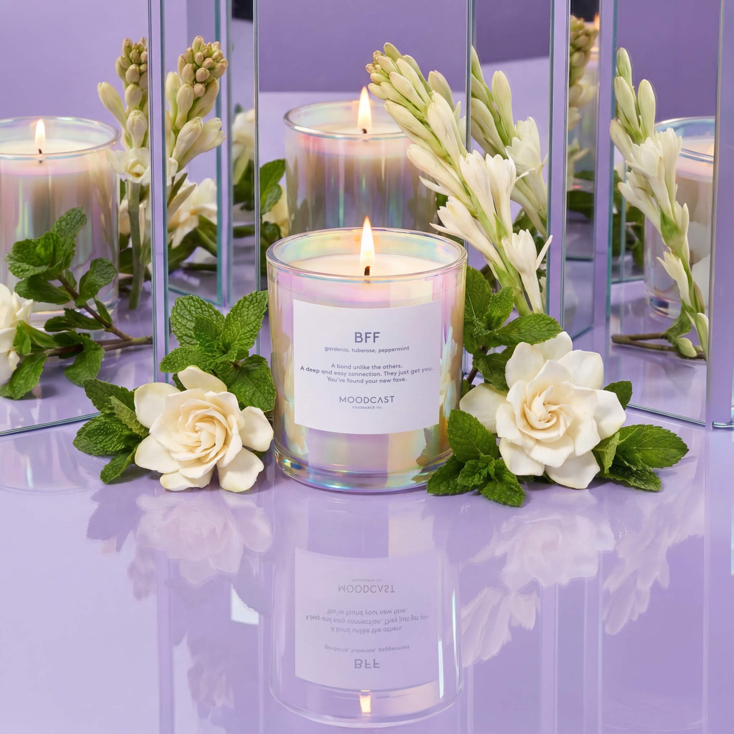 Lit candle in iridescent glass surrounded by white flowers and mint leaves on purple reflective surface