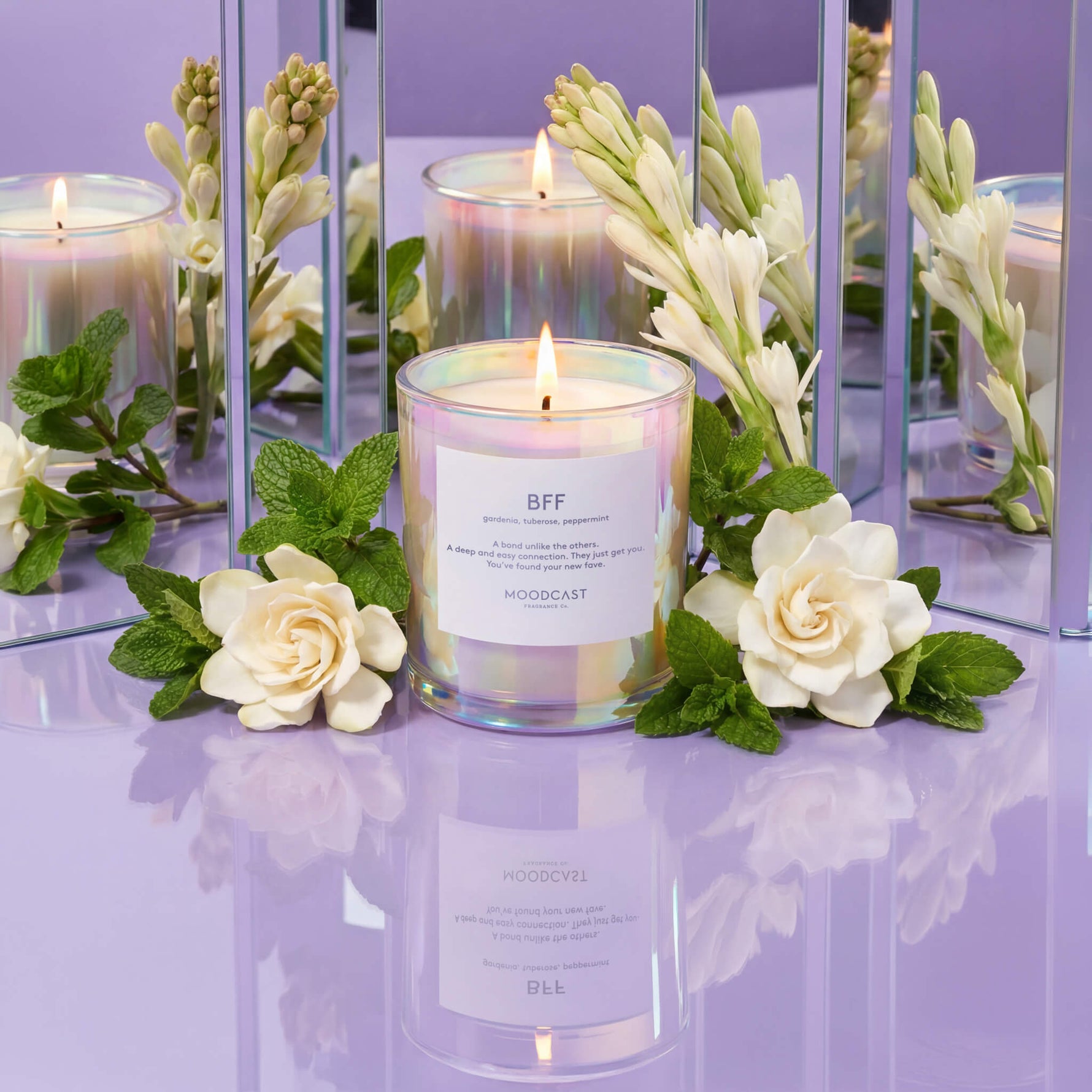 Lit candle in iridescent glass surrounded by white flowers and mint leaves on purple reflective surface