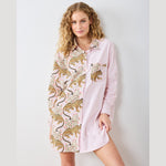 A smiling woman with long blonde curly hair models a unique split-design shirt dress against a white wall, featuring one side with pink and white vertical stripes and the other with a playful pale pink leopard and floral print