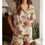 A woman stands indoors wearing a light-colored short-sleeved pajama set featuring golden leopards amidst green vines and pink flowers with a wooden cabinet and snake plant in the soft-focus background