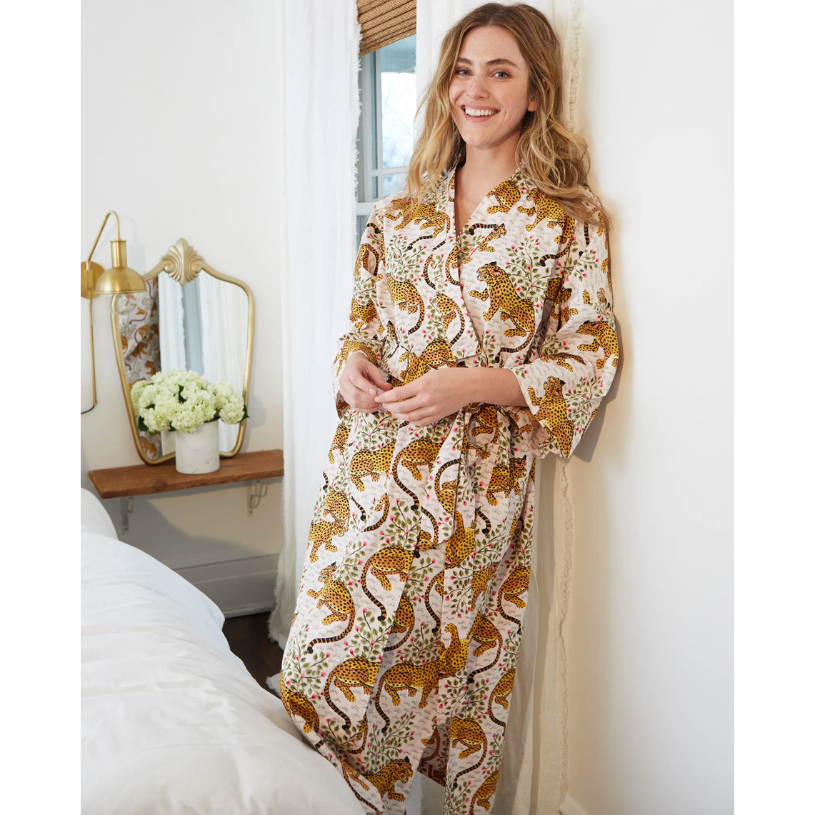 Smiling woman in a vibrant yellow leopard print robe with pink flowers and green leaves in a bright bedroom featuring a gold ornate mirror and white hydrangeas