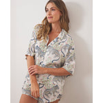 A woman with long blonde hair leans against a white wall, wearing a matching short-sleeved pajama set featuring blue and black spotted leopards intertwined with delicate pink and green floral motifs