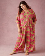 A smiling woman with dark wavy hair against a plain pink studio background wearing a vibrant pink two-piece loungewear set featuring striking yellow leopard prints and intricate green foliage motifs with one hand casually in a pocket