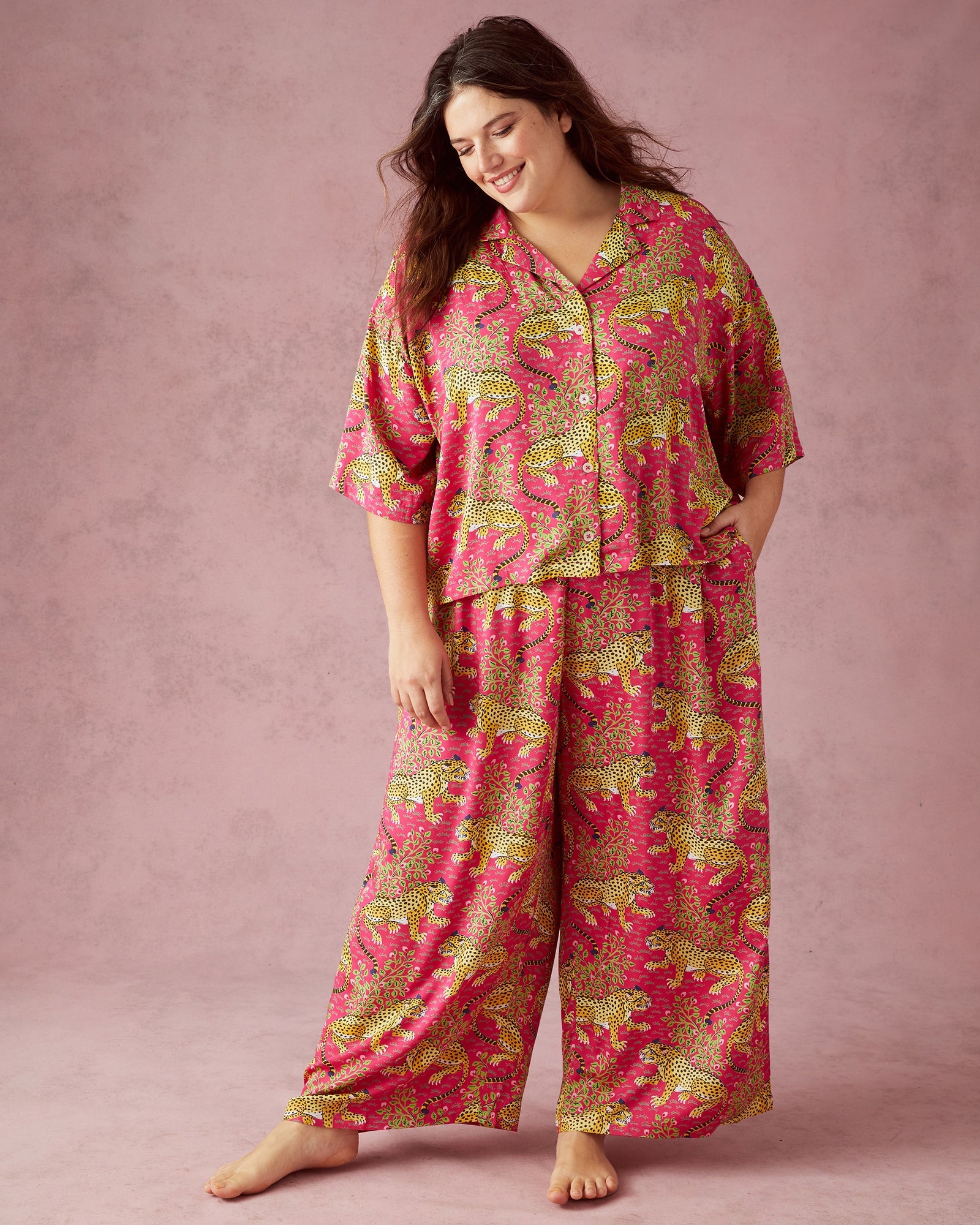 A smiling woman with dark wavy hair against a plain pink studio background wearing a vibrant pink two-piece loungewear set featuring striking yellow leopard prints and intricate green foliage motifs with one hand casually in a pocket