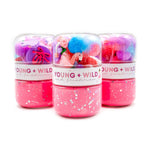 Product photography showing three Young + Wild and Friedman brand jars of vibrant pink glitter slime, topped with clear domes containing colorful feathers, a pink flamingo, and purple ballet slippers