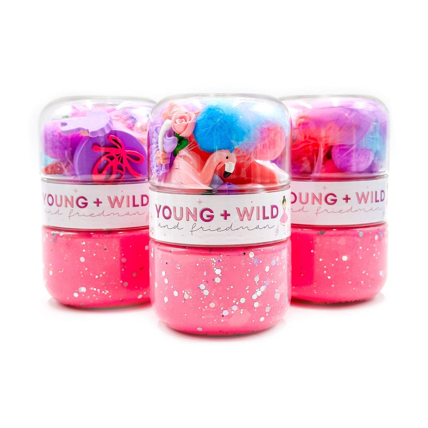 Product photography showing three Young + Wild and Friedman brand jars of vibrant pink glitter slime, topped with clear domes containing colorful feathers, a pink flamingo, and purple ballet slippers