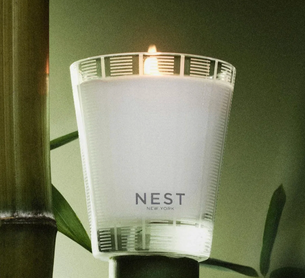 A lit NEST New York frosted glass candle with a unique banded pattern glows softly beside vibrant green bamboo stalks and leaves