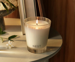 A lit NEST New York candle with a patterned glass holder on a light round table, beside a delicate white flower, green leaves, and stacked books bathed in soft warm light