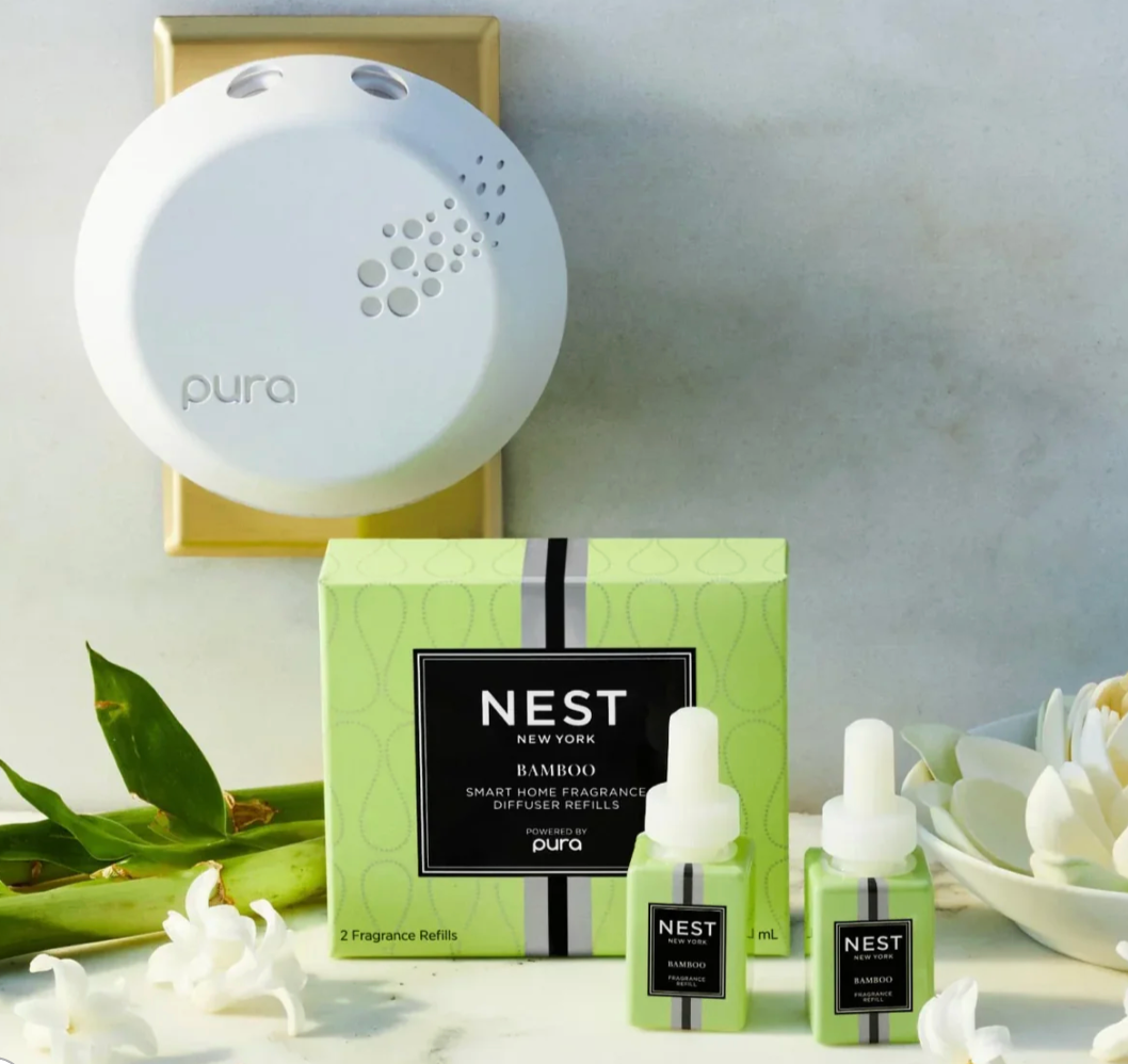 A white Pura smart home fragrance diffuser mounted on a gold wall plate above a vibrant green Nest New York Bamboo refill box, two small matching bottles, fresh green bamboo leaves, and delicate white flowers on a light marble surface