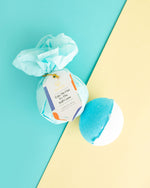 Musee bath bombs studio shot featuring one wrapped in light blue tissue paper with a Take Me Out To The Ball Game tag and one unwrapped half blue half white bath bomb on a diagonal light blue and yellow background