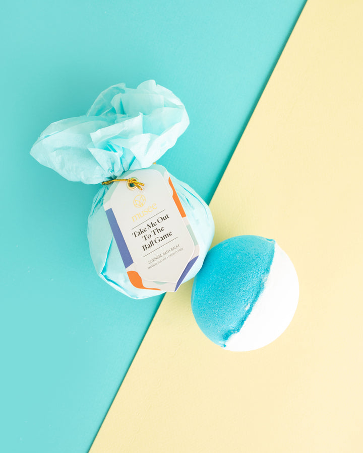 Musee bath bombs studio shot featuring one wrapped in light blue tissue paper with a Take Me Out To The Ball Game tag and one unwrapped half blue half white bath bomb on a diagonal light blue and yellow background