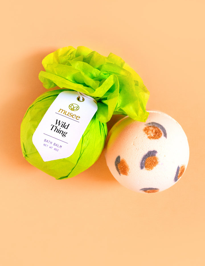 Digital photograph of Musee bath balms featuring one wrapped in vibrant green tissue paper with a Wild Thing tag and another white one with brown and grey leopard print spots on a peach background