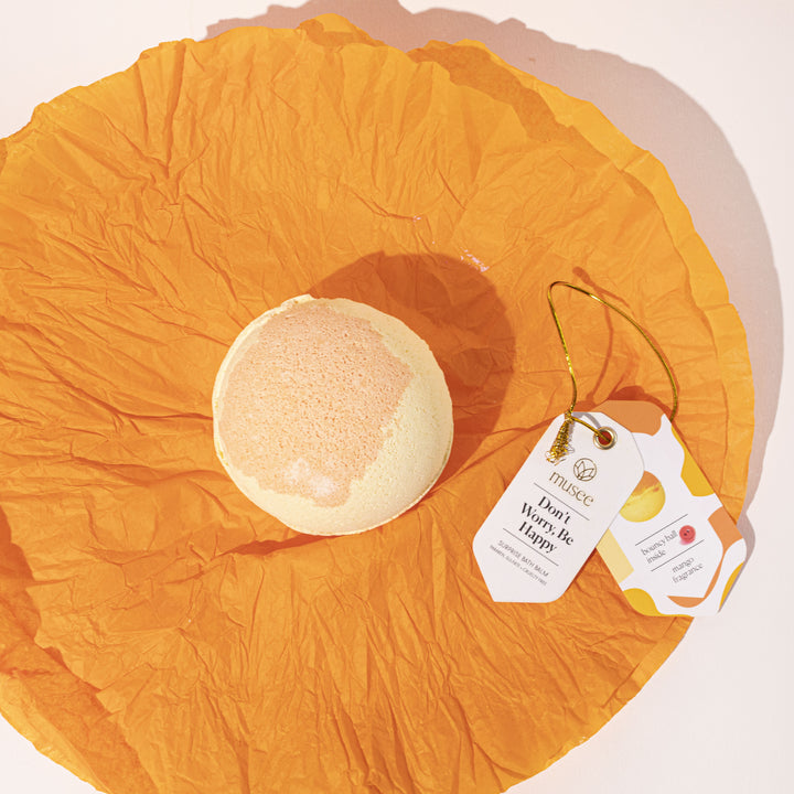 A pale yellow and peach Musee bath bomb rests on crumpled orange tissue paper next to two tags, one stating Don't Worry Be Happy and the other detailing bouncy ball inside with mango fragrance