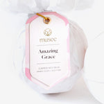 Photograph of a Musee Amazing Grace surprise bath balm in white tissue paper, featuring a pink and white patterned hang tag with golden 'Musee' text and eyelet