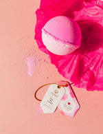 Photography of a two-toned pink and white Musee bath bomb resting on vibrant fuchsia tissue paper next to two white tags indicating 'You Are Loved' and 'heart inside sunflower fragrance' on a light pink surface