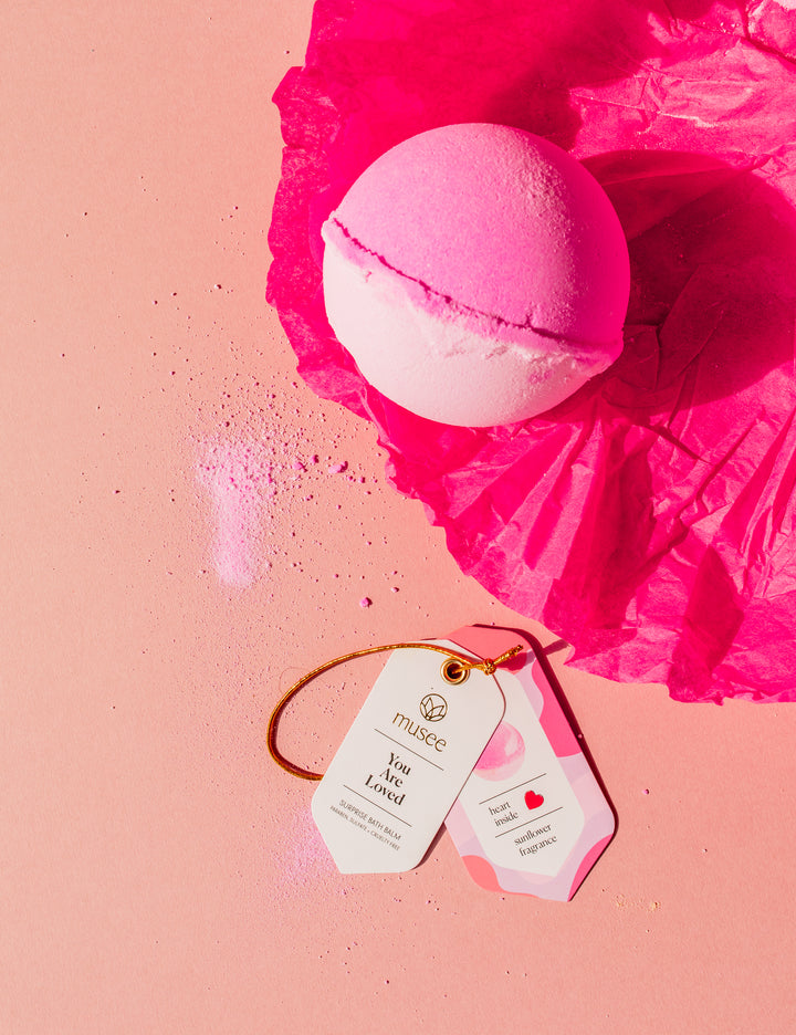 Photography of a two-toned pink and white Musee bath bomb resting on vibrant fuchsia tissue paper next to two white tags indicating 'You Are Loved' and 'heart inside sunflower fragrance' on a light pink surface