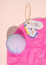 Musee's vibrant lavender and light blue swirled bath bomb rests on bright crumpled hot pink tissue paper next to its white product tag with a golden loop, featuring 'Let The Good Times Roll' and mentioning a surprise keychain inside with magnolia fragrance on a soft pink studio background