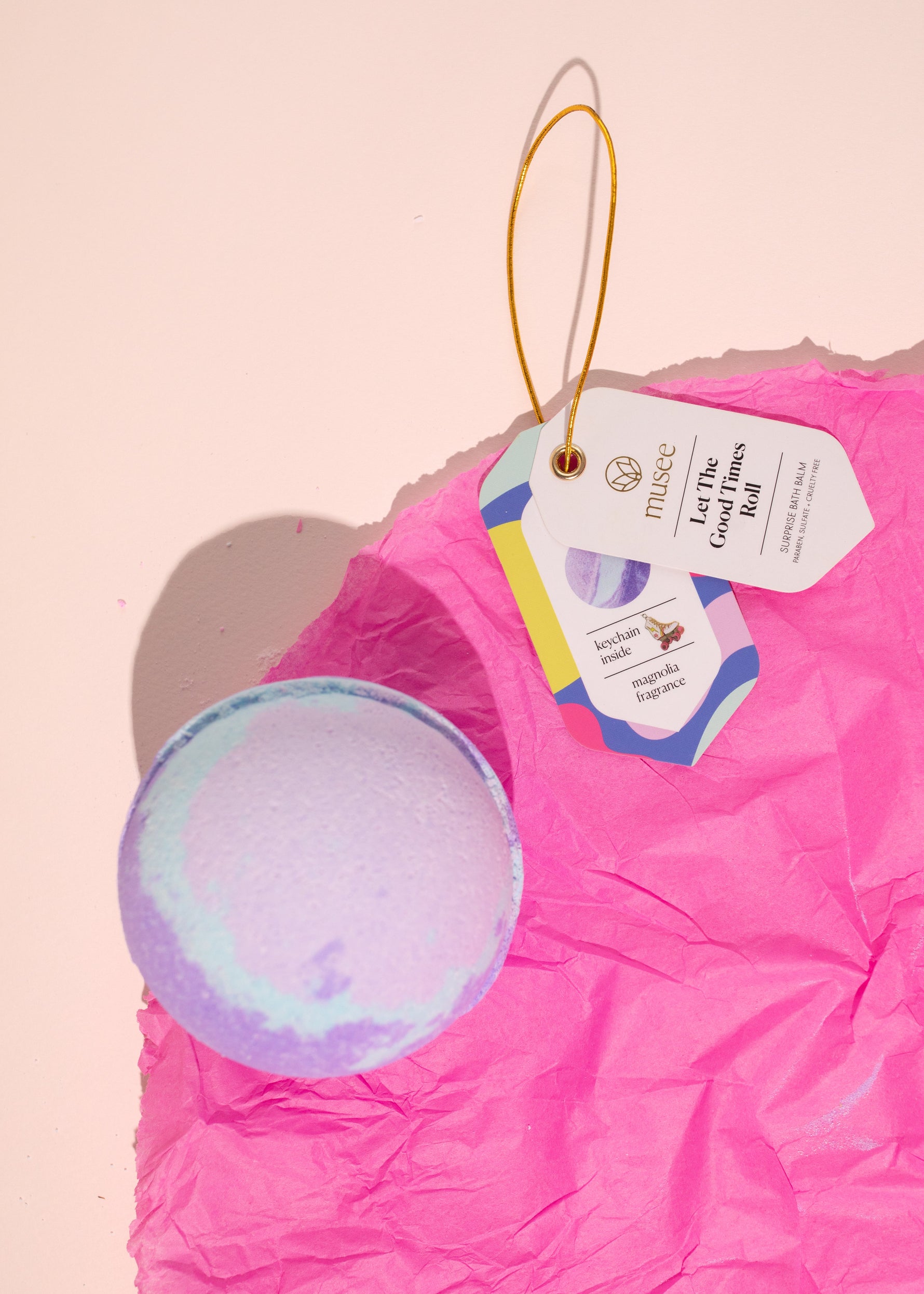 Musee's vibrant lavender and light blue swirled bath bomb rests on bright crumpled hot pink tissue paper next to its white product tag with a golden loop, featuring 'Let The Good Times Roll' and mentioning a surprise keychain inside with magnolia fragrance on a soft pink studio background