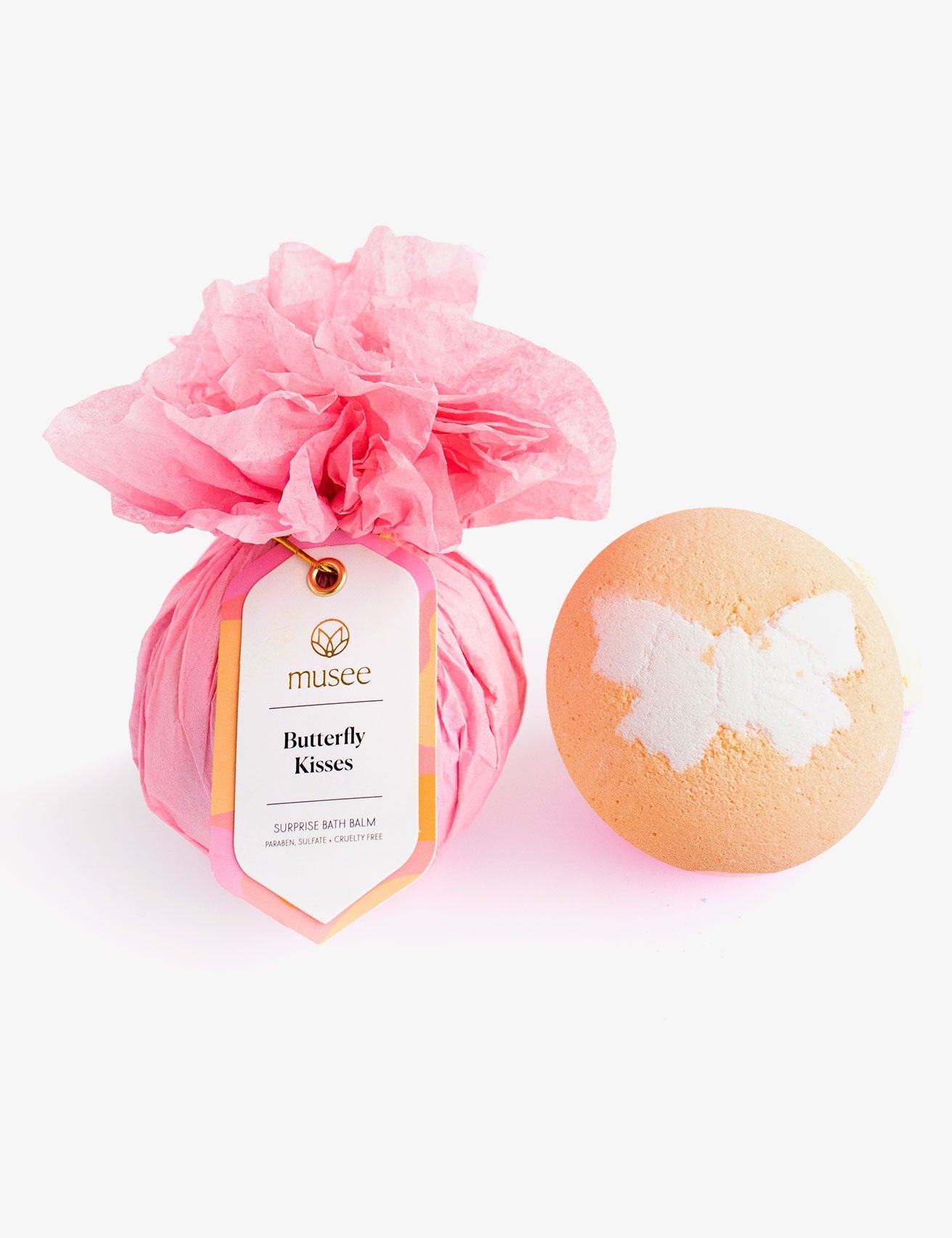 Product photograph by Musee showcasing two bath bombs on a white background one wrapped in pink tissue paper with a 'Butterfly Kisses Surprise Bath Balm' tag and another orange bath bomb featuring a white butterfly design