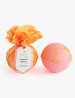 Photograph featuring a Musee Dancing Queen bath balm wrapped in vibrant orange tissue paper with a pink tag, alongside an unwrapped round pink bath bomb speckled with gold, all presented on a crisp white background