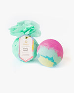 Product photo featuring two Musee bath bombs on a white background, one wrapped in mint green tissue paper with a "Feeling Groovy" tag, the other an unwrapped vibrant swirl of pink, light blue, and yellow