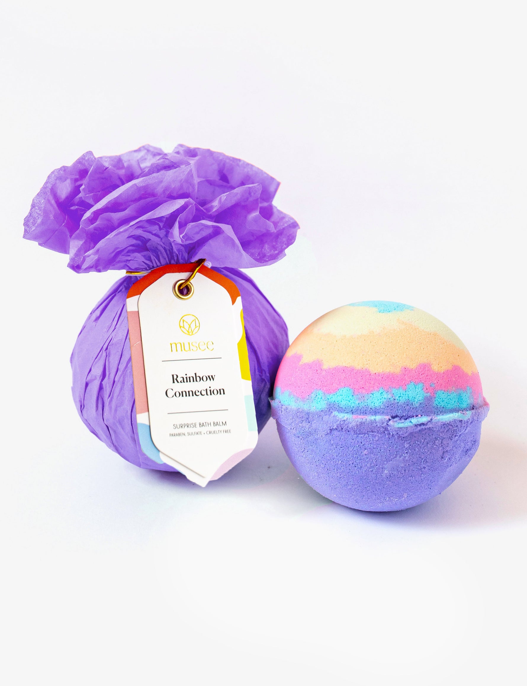Product photography by Musee featuring a bath balm wrapped in purple tissue paper with a "Rainbow Connection" tag next to a colorful unwrapped bath bomb displaying vibrant purple, blue, pink, and yellow layers on a white background
