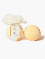 Product photography of two spherical bath balms by Musee on a white studio background one wrapped in crinkled white tissue with a gold-accented tag reading Tupelo Honey Surprise Bath Balm and the other unwrapped pale yellow