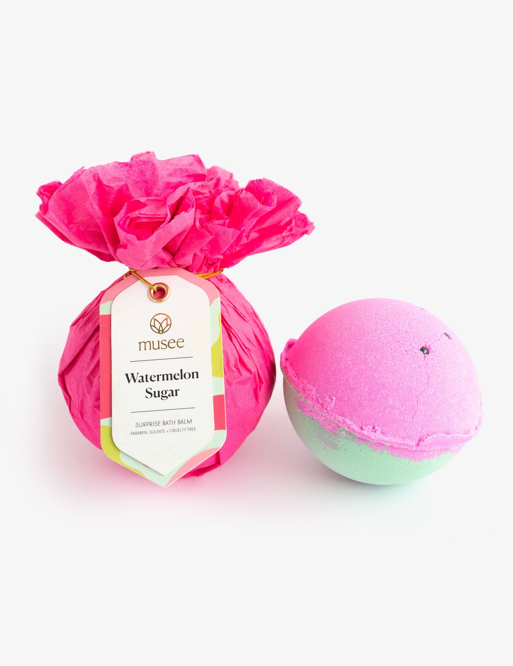 Product photo of a Musee Watermelon Sugar bath balm, one wrapped in bright pink tissue paper with a colorful brand tag, and a second unwrapped pink and light green bath bomb with dark speckles, on a clean white studio background