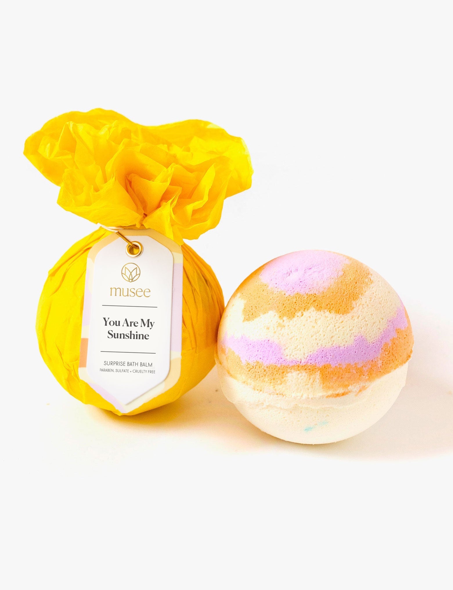 Product photography of two Musee bath bombs on a white background one wrapped in bright yellow tissue paper with a 'You Are My Sunshine Surprise Bath Balm' tag and the second showcasing swirls of white orange and lavender
