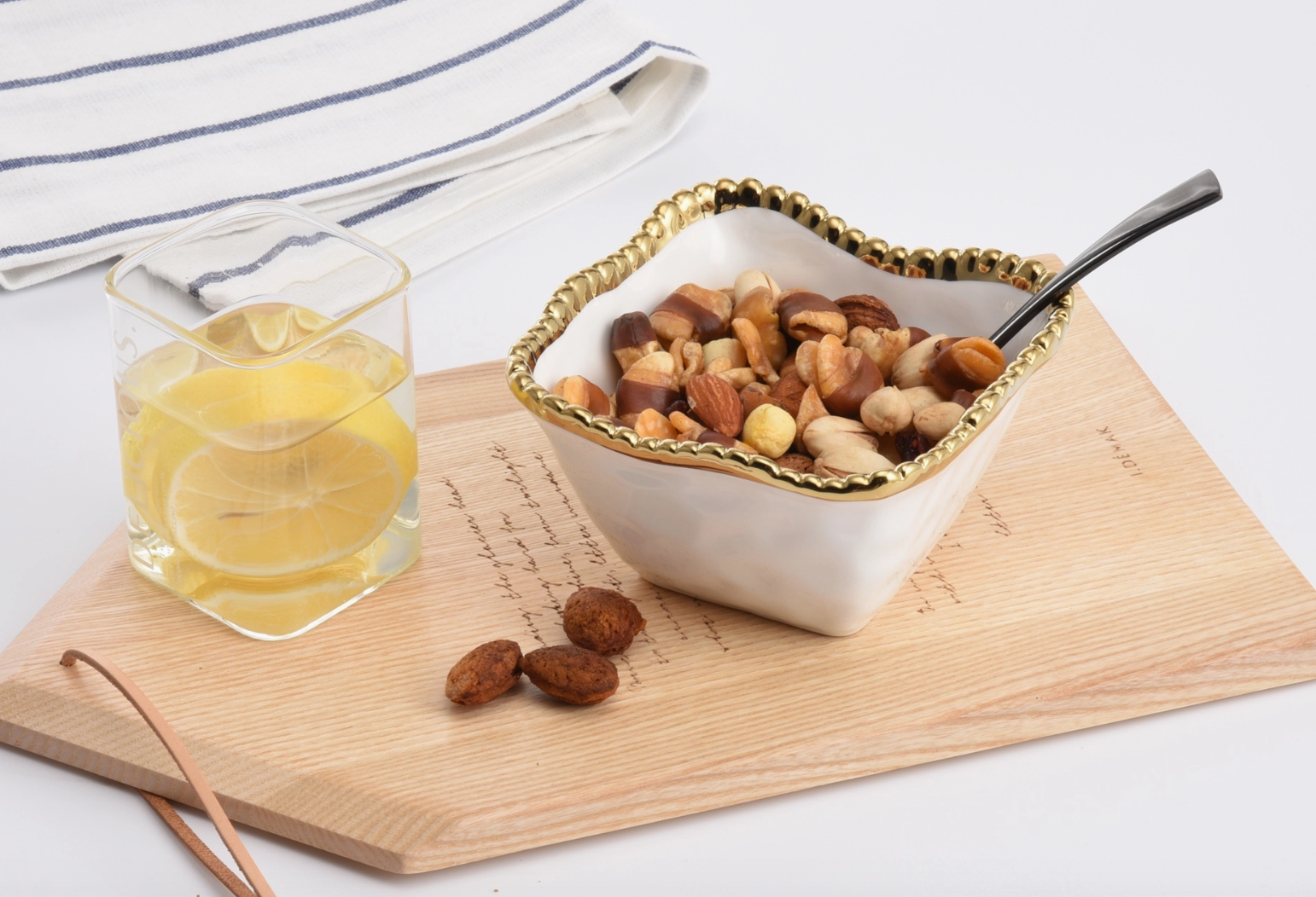 A white gold-rimmed bowl of assorted nuts with a dark spoon and a square glass of lemon water rest on a wooden board with decorative script beside a blue striped napkin