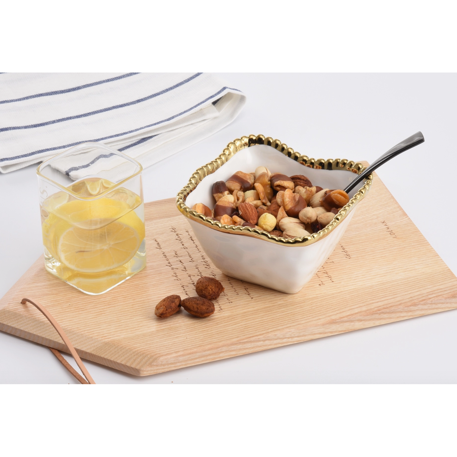 Still life photograph of a scalloped white bowl with a gold-beaded rim filled with mixed nuts resting on a wooden board with cursive script beside a square glass of lemon water and a blue-striped linen napkin